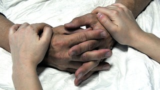 Hände eines älteren Menschen liegen gefaltet auf einer weißen Bettdecke und werden von einem jüngeren Menschen gehalten.