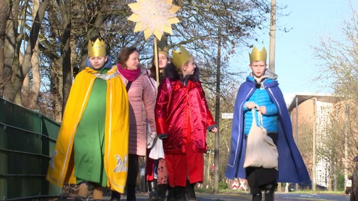 Mehrere Kinder sind als die drei Heiligen Könige bzw. als Sternsinger verkleidet und laufen mit Kronen auf dem Kopf die Straße entlang.