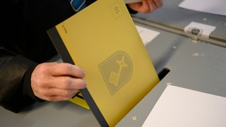 Eine Hand steckt einen Wahlzettel in eine Wahlurne.