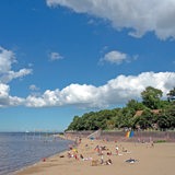 Links das Meer, rechts ein Sandstrand, mehrere Menschen liegen dort auf Strandhandtüchern.