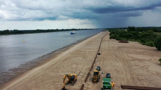 Bagger und Rohre sind an einem Strand an der Weser zu sehen.