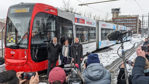 Drei Menschen stehen vor einer Straßenbahn und lächeln in Kameras von Journalisten.