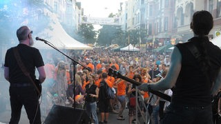 Auf einer Bühne stehen Männer mit Gitarren, davor tanzen Menschenmassen.