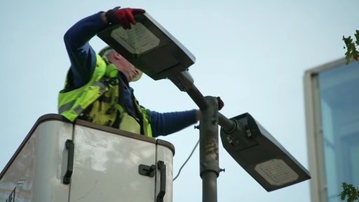 Von einem Bauarbeiter wird eine neue Lampe an eine Straßenlaterne angebracht.