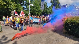 Verdi-Mitglieder laufen protestierend durch eine Bremer Straße. Sie sind umhüllt von roten und blauen Rauchschwaden.