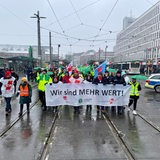 Mehrere Menschen laufen über Straßenbahn-Gleise vor dem Bremer Hauptbahnhof. Vor sich her tragen sie ein Transparent mit der Aufschrift "Wir sind MEHR WERT!"