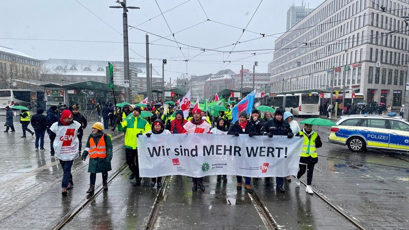 Mehrere Menschen laufen über Straßenbahn-Gleise vor dem Bremer Hauptbahnhof. Vor sich her tragen sie ein Transparent mit der Aufschrift "Wir sind MEHR WERT!"