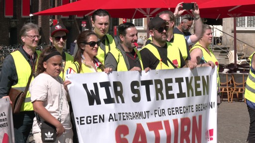 Es sind mehrere Personen mit gelben Warnwesten zu sehen, welche ein großes Plakat halten. Auf dem Plakat steht "Wir streiken".