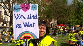 Eine Frau in einer gelben Warnweste hält ein Schild auf einer Kundgebung.