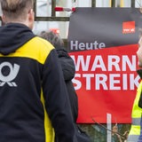 Auf einem Plakat steht "Heute Warnstreik". Davor stehen ein Mann in Briefzusteller-Kleidung und einer in Warnweste.
