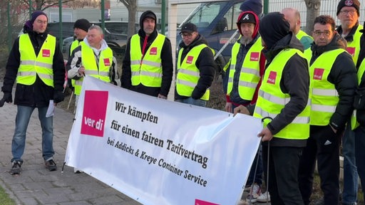 Mehrere Mitarbeitende bei Addicks & Kreye streiken, sie tragen gelbe Warnwesten und tragen einen Verdi Banner