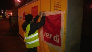 Ein Beschäftigter des Postzentrums Bremen hängt ein Verdi Plakat an eine Paketstation. 
