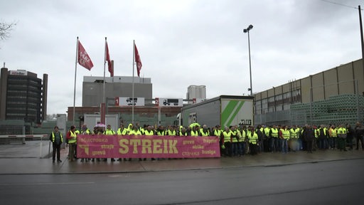 Mehrere Menschen stehen in gelben Warnwesten auf der Straße und halten ein Banner mit der Aufschrift "Streik" in den Händen.
