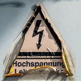 Schleswig, ein ausgeblichener und teils schon abgeloester Aufkleber mit einem Blitz-Piktogramm und der Aufschrift Hochspannung Lebensgefahr sowie Klebereste an einem Stromkasten.