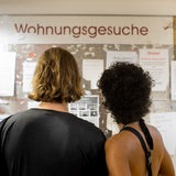 Zwei Frauen schauen auf ein Schwarzes Brett mit Wohnungsgesuchen. 