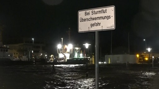 Ein Schild, auf dem "Bei Sturmflut Überschwemmungsgefahr" steht. Im Hintergrund die Überschwämmung.