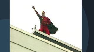 Ein Mann im roten Superheldenkostüm hebt die Faust während er auf einem Dach steht.