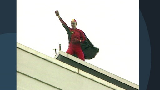 Ein Mann im roten Superheldenkostüm hebt die Faust während er auf einem Dach steht.
