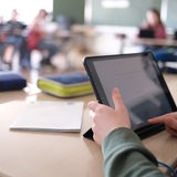 Ein Kind sitzt in einem Klassenzimmer. Vor sich hat es ein Tablet. Im Hintergrund sind weitere Kinder zu sehen.