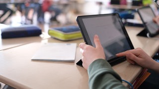 Ein Kind sitzt in einem Klassenzimmer. Vor sich hat es ein Tablet. Im Hintergrund sind weitere Kinder zu sehen.