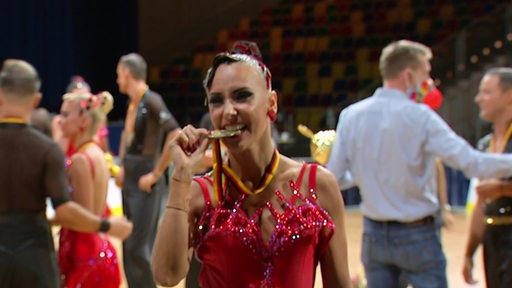 Eine Tänzerin beißt auf eine Gold-Medaillie und steht in einer Halle.