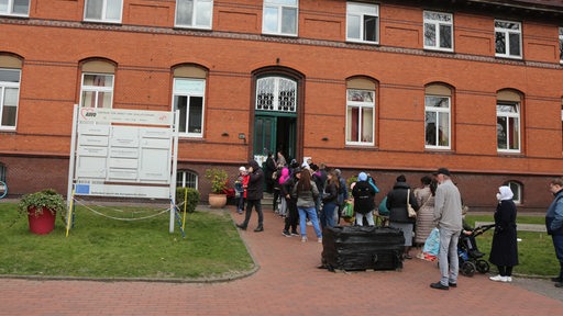 Menschen stehen in einer Schlange vor einem roten Backsteingebäude.