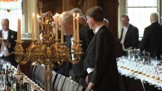 Ein Kerzenständer, der auf einer gedeckten Tafel steht. Im Hintergrund Männer in Anzügen.