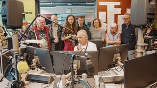 Eine Gruppe an Menschen steht hinter einem Radio-Moderator, der vor einem Mikrofon sitzt.