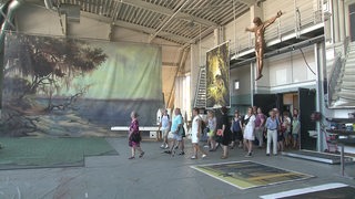 Menschen stehen in einer Werkhalle des Bremer Theaters. Über ihnen hängt eine Jesus-Figur.