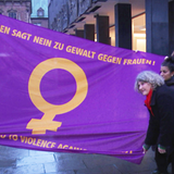 Mehrere Frauen halten eine Flagge, auf der Gewalt gegen Frauen verurteilt wird.