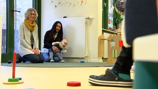 Zwei Frauen und ein Baby knien in einer Kindertagesstätte auf einem Teppich.