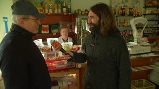 Freddy Radeke ist zu Besuch in einem Tante Emma Laden und redet mit einem Mann, während eine Frau hinter dem Verkaufstresen steht und die beiden Männer beobachtet.