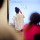 Ein Lehrerin schreibt eine Mathematikaufgabe auf eine digitale Schultafel im Klassenraum, im Vordergrund sitzen Schüler (Symbolbild)