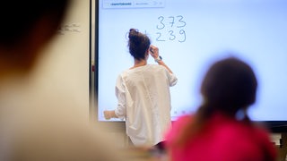 Ein Lehrerin schreibt eine Mathematikaufgabe auf eine digitale Schultafel im Klassenraum, im Vordergrund sitzen Schüler (Symbolbild)