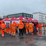Eine Gruppe Bauarbeiter in oranger Kleidung steht auf einem Hof.