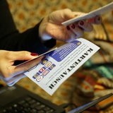 Eine Person mit rot lackierten Fingernägeln hält einen Flyer, auf dem "Kartentausch" steht, vor ihr steht ein Laptop