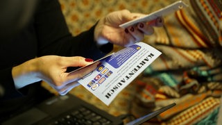 Eine Person mit rot lackierten Fingernägeln hält einen Flyer, auf dem "Kartentausch" steht, vor ihr steht ein Laptop