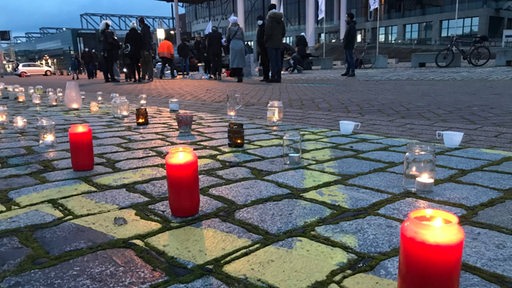 Auf der Bürgerweide stehen Kerzen auf dem Boden vor einer Halle. Im Hintergrund stehen Menschen.