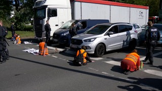 Mehrere Menschen sitzen in Warnwesten vor mehreren Autos auf einer Straße.