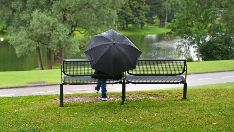 Eine Person sitzt mit Regenschirm auf einer Bank in einem Park (Archivbild)