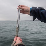 Zwei Personen halten ein Thermometer vor offenem Meer.