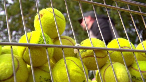 Ein Korb voller gelber Tennisbälle. Dahinter ein Mann mit Cappy im unscharfen Bereich.