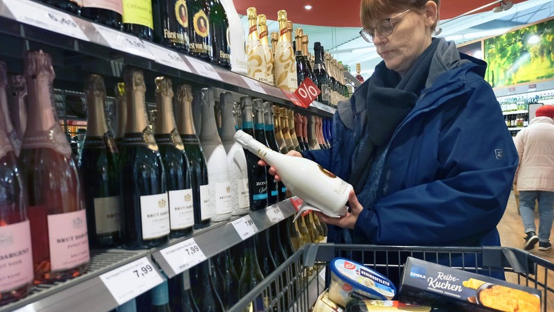 Eine Frau betrachtet im Supermarkt am Sektregal eine Flasche Sekt, im Einkaufswagen liegen diverse Festtagseinkäufe.