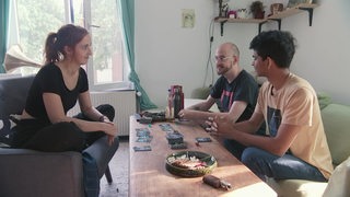 Die Bewohner einer WG, aus der Serie "Teuerland", sitzen am Wohnzimmertisch und spielen Karten.