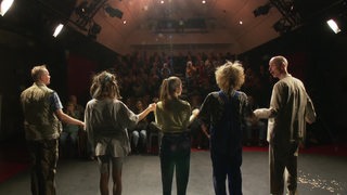 Fünf Schauspielerinnen und Schauspieler stehen Hand in Hand vor Publikum auf einer Bühne.