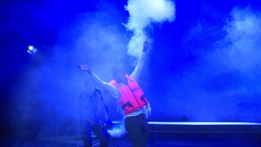 Es ist ein Mann in einer leuchtenden Rettungsweste zwischem weißem Nebel und blauen Licht zu sehen. 