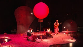 Die Inszenierung des Theaterstückes der Rote Baum, bei der drei Personen unter einem roten Ballon sitzen.