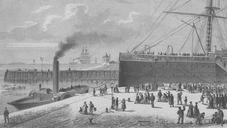 Ein Schwarz-Weiß-Bild zeigt eine Szene im Hafen, vorne liegt ein Dampfer, hinten ein großes Schiff, drumherum viele Menschen.