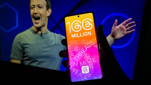 Ein Smartphone liegt in einer Hand. Darauf zu sehen der Text 100 Millionen Threads. Im Hintergrund ein Mann, der aussieht als würde er sich freuen.