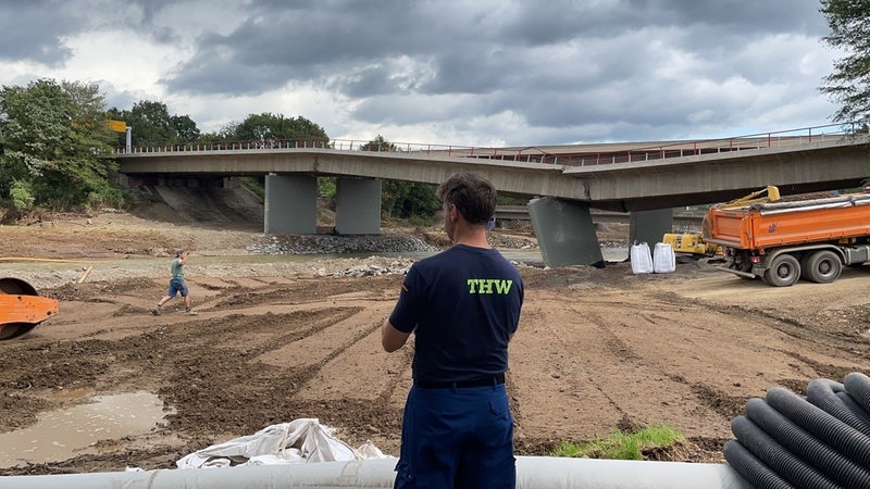 Ein Mann blickt auf zerstörtes Gelände und eine kaputte Flussbrücke.